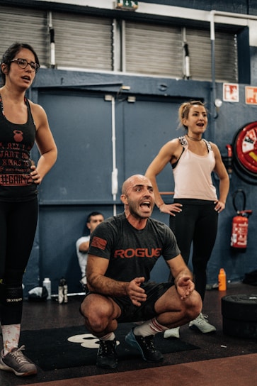 A group of people in an indoor gym setting, with two women standing and a man squatting and appearing to be shouting encouragement. They are dressed in workout clothes, surrounded by gym equipment such as weights and a red fire extinguisher in the background.