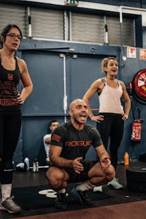 A group of people in an indoor gym setting, with two women standing and a man squatting and appearing to be shouting encouragement. They are dressed in workout clothes, surrounded by gym equipment such as weights and a red fire extinguisher in the background.