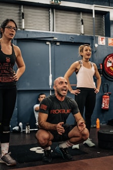 A group of people in an indoor gym setting, with two women standing and a man squatting and appearing to be shouting encouragement. They are dressed in workout clothes, surrounded by gym equipment such as weights and a red fire extinguisher in the background.