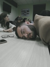 A group of people are seated around a table in what appears to be a meeting or classroom setting. One person has their head resting on the table, seemingly asleep. Eyeglasses and a phone are placed on the table nearby. The atmosphere appears to be calm but possibly indicative of boredom or fatigue.
