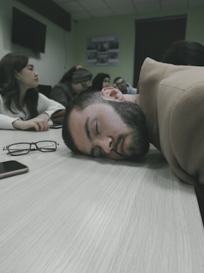 A group of people are seated around a table in what appears to be a meeting or classroom setting. One person has their head resting on the table, seemingly asleep. Eyeglasses and a phone are placed on the table nearby. The atmosphere appears to be calm but possibly indicative of boredom or fatigue.