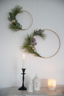 Two minimalist wreaths with pine and eucalyptus leaves are mounted on a white wall. They are adorned with golden ornaments. Below the wreaths, a single white candle stands on a black holder, accompanied by small white ceramic houses and a lit white candle in a glass holder, creating a cozy and festive atmosphere.