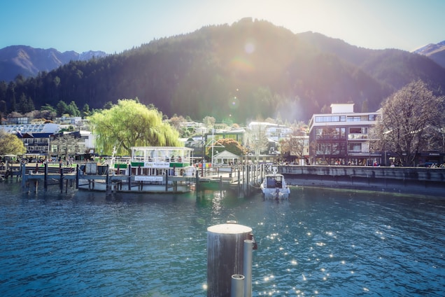 A serene waterfront scene with a dock featuring several boats and a floating bar. In the background, there are modern buildings and lush trees with a mountainous landscape rising behind them. The sun is casting a warm glow over the area, creating a peaceful and picturesque setting.