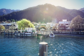 A serene waterfront scene with a dock featuring several boats and a floating bar. In the background, there are modern buildings and lush trees with a mountainous landscape rising behind them. The sun is casting a warm glow over the area, creating a peaceful and picturesque setting.