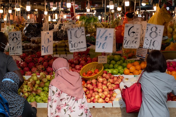 A bustling fruit market with a variety of colorful fruits neatly arranged. Several handwritten signs indicate prices and offers. Numerous shoppers, some wearing headscarves and masks, are browsing and examining the fresh produce. The lighting is warm, with numerous light bulbs hanging from the ceiling.