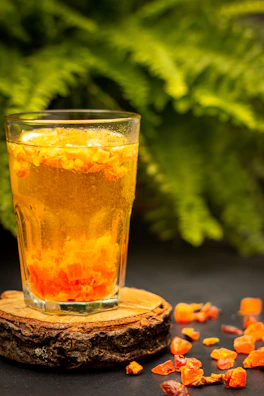 A glass filled with an orange and yellow drink containing pieces of fruit at the bottom is placed on a wooden coaster. Dried orange fruit pieces are scattered around on a dark surface. A backdrop of lush green ferns creates a vibrant contrast.