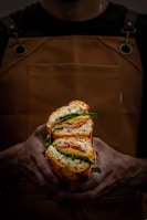A person wearing a dark shirt and brown apron is holding a large sandwich with both hands. The sandwich is cut in half, revealing layers of lettuce, sliced meats, and cheese, all within a crusty bread.