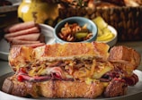 A gourmet sandwich filled with caramelized onions, thin slices of cured meat, and melted cheese between crispy toasted bread. In the background, there are slices of what looks like other meats on a different plate, a small bowl of pickles, and a woven basket containing more bread or rolls. The setup is arranged on a tabletop, suggesting a rustic or artisanal dining experience.