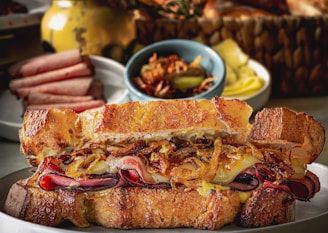 A gourmet sandwich filled with caramelized onions, thin slices of cured meat, and melted cheese between crispy toasted bread. In the background, there are slices of what looks like other meats on a different plate, a small bowl of pickles, and a woven basket containing more bread or rolls. The setup is arranged on a tabletop, suggesting a rustic or artisanal dining experience.
