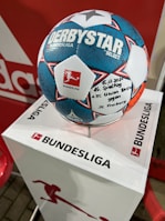 A soccer ball with the text DERBYSTAR and BUNDESLIGA is placed on a white pedestal that also has the BUNDESLIGA logo. The ball features a distinctive blue, white, and red pattern, with handwritten text noting the date 15.12.2021 with a mention of a match involving 1. FC Union Berlin and SC Freiburg.