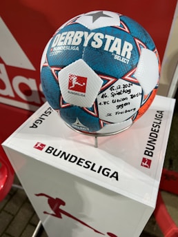 A soccer ball with the text DERBYSTAR and BUNDESLIGA is placed on a white pedestal that also has the BUNDESLIGA logo. The ball features a distinctive blue, white, and red pattern, with handwritten text noting the date 15.12.2021 with a mention of a match involving 1. FC Union Berlin and SC Freiburg.