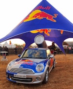 A branded Mini Cooper car with a large Red Bull can mounted on top is parked under a blue and red canopy featuring the Red Bull logo. The scene appears to be an outdoor event with several people in the background and other event setups visible.