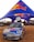 A branded Mini Cooper car with a large Red Bull can mounted on top is parked under a blue and red canopy featuring the Red Bull logo. The scene appears to be an outdoor event with several people in the background and other event setups visible.