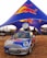 A branded Mini Cooper car with a large Red Bull can mounted on top is parked under a blue and red canopy featuring the Red Bull logo. The scene appears to be an outdoor event with several people in the background and other event setups visible.