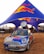 A branded Mini Cooper car with a large Red Bull can mounted on top is parked under a blue and red canopy featuring the Red Bull logo. The scene appears to be an outdoor event with several people in the background and other event setups visible.