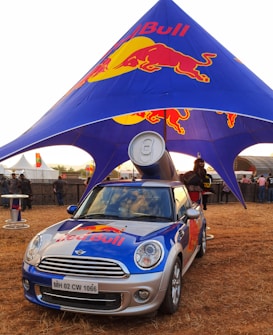 A branded Mini Cooper car with a large Red Bull can mounted on top is parked under a blue and red canopy featuring the Red Bull logo. The scene appears to be an outdoor event with several people in the background and other event setups visible.