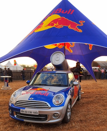 A branded Mini Cooper car with a large Red Bull can mounted on top is parked under a blue and red canopy featuring the Red Bull logo. The scene appears to be an outdoor event with several people in the background and other event setups visible.
