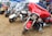 A gathering of several colorful motorcycles parked on dry grass, each with distinctive designs and features, situated outdoors under a blue tent. The motorcycles are lined up side by side, showcasing various models, including one with a striking red and black color scheme.