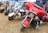 A gathering of several colorful motorcycles parked on dry grass, each with distinctive designs and features, situated outdoors under a blue tent. The motorcycles are lined up side by side, showcasing various models, including one with a striking red and black color scheme.