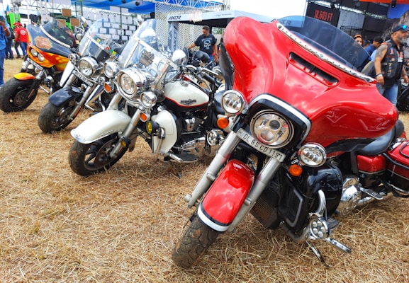 A gathering of several colorful motorcycles parked on dry grass, each with distinctive designs and features, situated outdoors under a blue tent. The motorcycles are lined up side by side, showcasing various models, including one with a striking red and black color scheme.