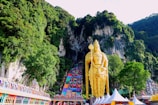 A towering golden statue stands prominently against a background of lush green hills and rugged cliffs. Colorful steps lead up to a temple, with people ascending. The base of the statue and steps features ornate carvings and vibrant decorations. The scene is vibrant and lively, capturing both natural beauty and cultural richness.