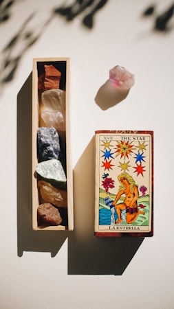A tarot card, labeled 'La Estrella', depicting a figure pouring water is placed next to a wooden box containing various crystals of different colors. A separate pink crystal is positioned nearby, casting a shadow.