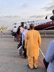 A group of people standing in line, waiting to board an airplane on a tarmac. The aircraft is white with maroon accents and displays the logo of Arik Air. Some individuals are carrying bags and dressed in a variety of attire.