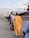 A group of people standing in line, waiting to board an airplane on a tarmac. The aircraft is white with maroon accents and displays the logo of Arik Air. Some individuals are carrying bags and dressed in a variety of attire.