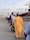 A group of people standing in line, waiting to board an airplane on a tarmac. The aircraft is white with maroon accents and displays the logo of Arik Air. Some individuals are carrying bags and dressed in a variety of attire.