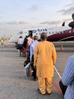 A group of people standing in line, waiting to board an airplane on a tarmac. The aircraft is white with maroon accents and displays the logo of Arik Air. Some individuals are carrying bags and dressed in a variety of attire.