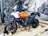 A motorcycle with an orange and white tank is displayed on a wooden platform. The setting appears to be an indoor or semi-outdoor exhibition with rocks caged behind the vehicle and a large mural depicting bikers on an open road. The motorcycle features sturdy tires and a prominent windshield, suggesting it is designed for adventure or off-road travel.