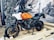 A motorcycle with an orange and white tank is displayed on a wooden platform. The setting appears to be an indoor or semi-outdoor exhibition with rocks caged behind the vehicle and a large mural depicting bikers on an open road. The motorcycle features sturdy tires and a prominent windshield, suggesting it is designed for adventure or off-road travel.