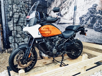 A motorcycle with an orange and white tank is displayed on a wooden platform. The setting appears to be an indoor or semi-outdoor exhibition with rocks caged behind the vehicle and a large mural depicting bikers on an open road. The motorcycle features sturdy tires and a prominent windshield, suggesting it is designed for adventure or off-road travel.