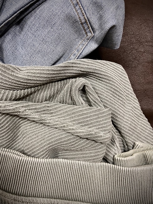 A close-up of fabrics with different textures. The top fabric is a light blue denim with visible stitching, while the lower section is covered with a ribbed knit fabric in a light grey color. These materials are layered on what appears to be a dark brown leather surface.