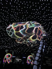 A large, abstract structure resembling a brain with colorful, swirling patterns is suspended in a room with a ceiling of glowing white stars. Below, illuminated multicolored loops or rings add to the vibrant atmosphere. The environment suggests an immersive, possibly digital or futuristic setting.