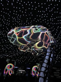 A large, abstract structure resembling a brain with colorful, swirling patterns is suspended in a room with a ceiling of glowing white stars. Below, illuminated multicolored loops or rings add to the vibrant atmosphere. The environment suggests an immersive, possibly digital or futuristic setting.