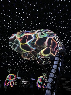 A large, abstract structure resembling a brain with colorful, swirling patterns is suspended in a room with a ceiling of glowing white stars. Below, illuminated multicolored loops or rings add to the vibrant atmosphere. The environment suggests an immersive, possibly digital or futuristic setting.
