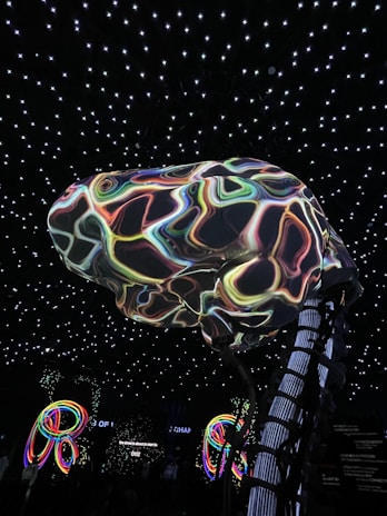 A large, abstract structure resembling a brain with colorful, swirling patterns is suspended in a room with a ceiling of glowing white stars. Below, illuminated multicolored loops or rings add to the vibrant atmosphere. The environment suggests an immersive, possibly digital or futuristic setting.