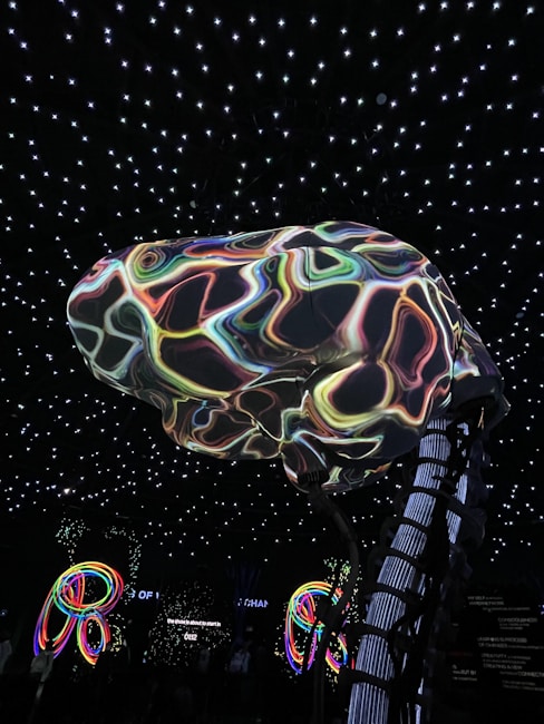 A large, abstract structure resembling a brain with colorful, swirling patterns is suspended in a room with a ceiling of glowing white stars. Below, illuminated multicolored loops or rings add to the vibrant atmosphere. The environment suggests an immersive, possibly digital or futuristic setting.