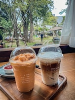 Two iced coffee beverages are placed on a wooden tray inside a cozy café setting. The left cup has a light brown, creamy appearance, topped with a dome lid featuring frothy toppings. The text on the cup reads 'IT'S OK NOT TO DRINK COFFEE #WITHIN'. The right cup shows a layered coffee with milk, also capped with a transparent dome lid. The text on this cup reads 'WITHIN THIS CUP' along with a milk layer illustration. In the background, an outdoor garden with trees and tables is visible, and a person is walking by.