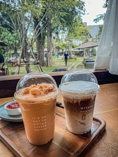 Two iced coffee beverages are placed on a wooden tray inside a cozy café setting. The left cup has a light brown, creamy appearance, topped with a dome lid featuring frothy toppings. The text on the cup reads 'IT'S OK NOT TO DRINK COFFEE #WITHIN'. The right cup shows a layered coffee with milk, also capped with a transparent dome lid. The text on this cup reads 'WITHIN THIS CUP' along with a milk layer illustration. In the background, an outdoor garden with trees and tables is visible, and a person is walking by.