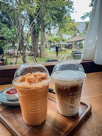Two iced coffee beverages are placed on a wooden tray inside a cozy café setting. The left cup has a light brown, creamy appearance, topped with a dome lid featuring frothy toppings. The text on the cup reads 'IT'S OK NOT TO DRINK COFFEE #WITHIN'. The right cup shows a layered coffee with milk, also capped with a transparent dome lid. The text on this cup reads 'WITHIN THIS CUP' along with a milk layer illustration. In the background, an outdoor garden with trees and tables is visible, and a person is walking by.