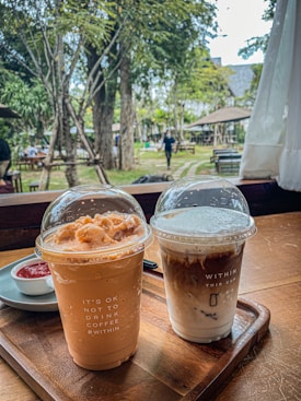 Two iced coffee beverages are placed on a wooden tray inside a cozy café setting. The left cup has a light brown, creamy appearance, topped with a dome lid featuring frothy toppings. The text on the cup reads 'IT'S OK NOT TO DRINK COFFEE #WITHIN'. The right cup shows a layered coffee with milk, also capped with a transparent dome lid. The text on this cup reads 'WITHIN THIS CUP' along with a milk layer illustration. In the background, an outdoor garden with trees and tables is visible, and a person is walking by.