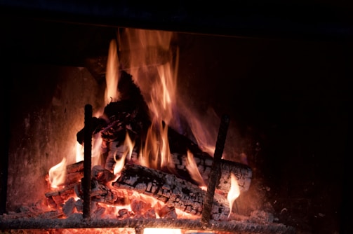 A close-up view of a burning fireplace with wooden logs, encased in a metallic structure. The flames are bright, flickering in shades of orange and yellow, illuminating the charred wood. Some glowing embers and ashes are present at the base.