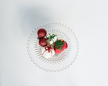 A clear glass dish holds two red glittery Christmas ornaments and three small decorative items resembling gift boxes and a Christmas bauble, all with festive colors and intricate designs.