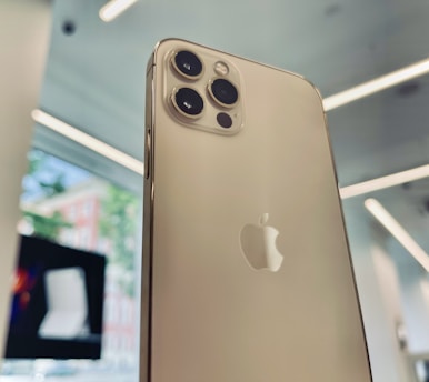 A close-up view of a modern smartphone with a metallic finish, featuring multiple camera lenses and a brand logo. The background shows an interior setting with bright lighting and blurred elements like a window and another electronic device.