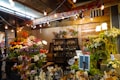 A vibrant flower shop with a wide variety of colorful blooms including sunflowers, lilies, and roses displayed on tables. The setting is warmly lit with hanging bulbs and an array of potted plants is arranged on shelves behind a counter. A person stands at the counter, partially hidden by the flowers.