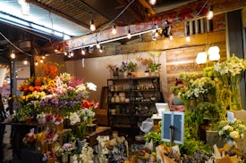 A vibrant flower shop with a wide variety of colorful blooms including sunflowers, lilies, and roses displayed on tables. The setting is warmly lit with hanging bulbs and an array of potted plants is arranged on shelves behind a counter. A person stands at the counter, partially hidden by the flowers.