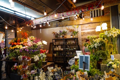 A vibrant flower shop with a wide variety of colorful blooms including sunflowers, lilies, and roses displayed on tables. The setting is warmly lit with hanging bulbs and an array of potted plants is arranged on shelves behind a counter. A person stands at the counter, partially hidden by the flowers.