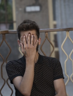 A person is seated with their hand covering their face, obscuring their identity. They have a tattoo on their wrist, and the background includes a patterned rail and a slightly blurred wall.